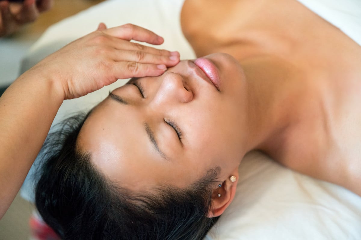 A close-up, overhead view of a woman with her eyes closed receiving a facial massage from a technician at a spa.