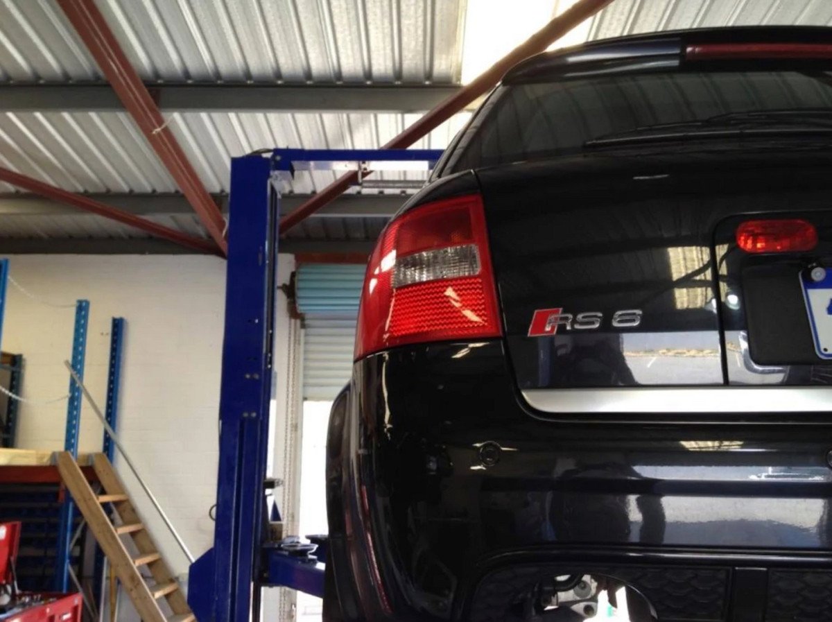 The rear left side of a black Audi RS 6, positioned on a blue hydraulic car lift inside a garage with a corrugated metal ceiling.