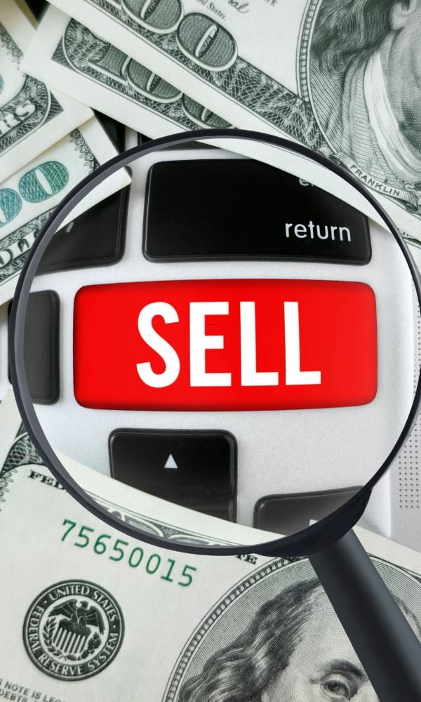 A magnifying glass focused on a red "SELL" button on a computer keyboard, resting on a pile of one-hundred-dollar bills.
