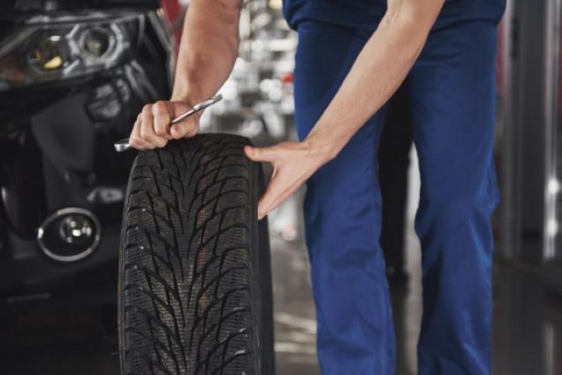 A close-up of a person in blue workwear holding a large, new vehicle tire with a deep tread pattern, while holding a wrench in their other hand.
