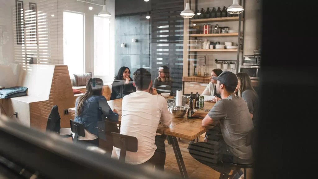 A diverse group of people sits around a long wooden communal table in a modern, sunlit breakroom or office kitchen.